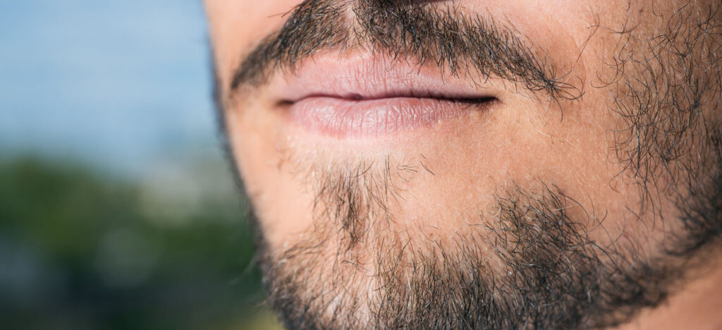 Zbliżenie na twarz mężczyzny z rzadkim zarostem, ukazujące nieregularny wzrost włosów na brodzie i wąsach